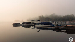 Moored boats reflected in still water at dawn with forested shoreline shrouded in fog.