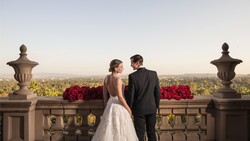 Bride and groom viewing expansive landscape from stone balustrade with ornamental urns and red floral arrangements.