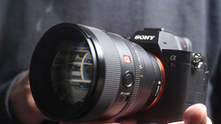 Sony mirrorless camera with telephoto lens mounted, held in hands against blurred gray background.