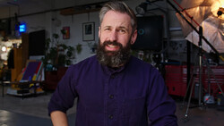 Bearded man in dark purple work shirt smiling in a workshop studio setting.