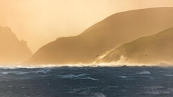 Dramatic coastal landscape with golden-hour light illuminating steep cliffs and turbulent ocean waves.