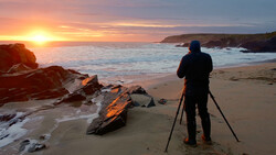 Photographer on a beach with tripod watching golden sunrise over ocean with rocky outcrops.