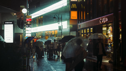 Rainy urban street at night with pedestrians holding umbrellas beneath illuminated storefronts and signs.
