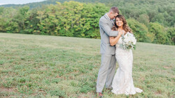 Bride and groom embracing in an open field with forested hills in the background.