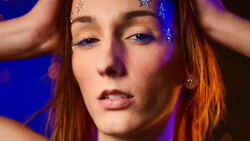 Woman with reddish-brown hair and blue eyeshadow against purple-blue lighting.