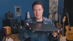 Man in a plaid shirt holding a camera lens in a home studio setup with guitars and audio equipment.