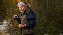 Photographer with gray hair holding a camera in an autumn forest setting.