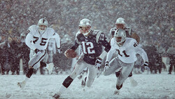 Four football players in a heavy snowstorm, two wearing white uniforms and two in dark uniforms with numbers visible.