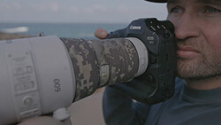 Photographer holding Canon EOS R1 with white telephoto lens featuring digital camouflage pattern.