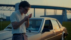 Man in glasses and white shirt eating a slice of pizza outdoors beside a vintage sedan.