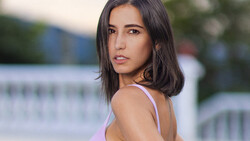 Woman with long dark hair wearing a lavender tank top, photographed outdoors near a waterfront.