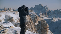 Photographer in black jacket holding camera to face on snowy mountain peak with dramatic rocky peaks in background.