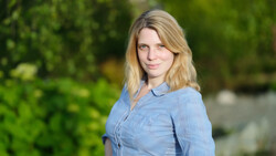 Woman with blonde shoulder-length hair wearing a light blue button-up shirt, photographed outdoors with soft natural lighting.
