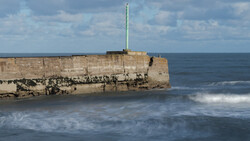 Weathered stone pier or breakwater extending into rough ocean waves under partly cloudy sky.