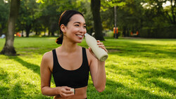 Athletic woman in black sports bra drinking from white water bottle in sunny park.