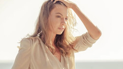 Woman in beige shirt with hand raised to head, backlit by golden sunlight against neutral background.