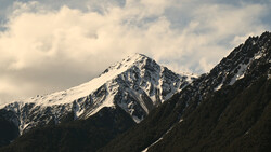 Snow-capped mountain peak with forested slopes under partly cloudy sky.