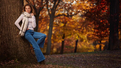 Woman leaning against tree trunk in autumn forest wearing cream cardigan and jeans.