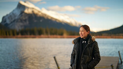 Woman in black quilted jacket stands at lake shore with snow-capped mountain in background.
