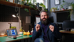 Bearded man in dark jacket sits at desk with laptop, gesturing while speaking in a plant-filled studio.