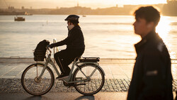 Person riding a bicycle on a waterfront promenade at golden hour with a blurred figure walking nearby.