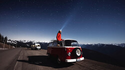 Person standing on roof of red and white vintage van at night, shining flashlight skyward with snow-capped mountains and starry sky in background.