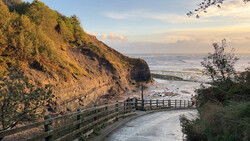 Wooden boardwalk descending a grass-covered cliff toward a calm sea at golden hour.