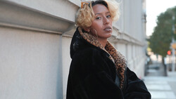 Woman with blonde curled hair wearing black velvet jacket leaning against white wall.