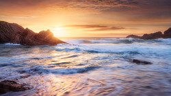Waves crash on a rocky beach at sunset, with golden light illuminating the water and headlands.