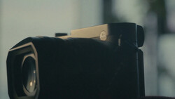 Black compact camera with a beige/tan hot shoe and visible lens, photographed against a soft blurred background.