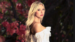 Woman with blonde wavy hair wearing an off-shoulder white ruffled top, photographed against a blurred background of pink flowering branches.