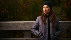 Woman in puffy jacket and black beanie sitting against wooden railing with blurred forest background.