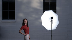 Woman in red shirt posing against gray brick wall lit by large octagonal softbox.