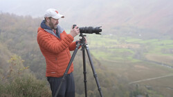Photographer with telephoto lens mounted on tripod, positioned on elevated terrain overlooking misty valley landscape.
