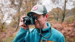 Photographer holding a camera to his face while wearing a turquoise fleece jacket and cap in an autumn landscape.