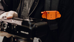 Photographer holding a camera with an orange camera strap accessory visible against dark clothing.