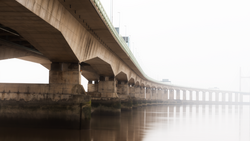 Long concrete bridge with supporting columns disappearing into thick fog over calm water.