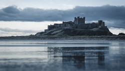 Medieval castle ruins on a rocky island reflected in still tidal flats under dramatic cloudy sky.