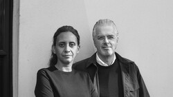 Two people standing against a white wall in a black and white portrait photograph.