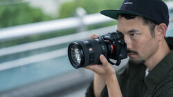 Photographer holding a mirrorless camera with a telephoto lens, wearing a black cap and olive shirt.