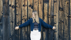Woman in navy blazer and white skirt posing with arms outstretched against weathered wooden backdrop.