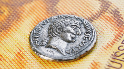 Ancient Roman coin with profile portrait resting on colorful euro banknotes.