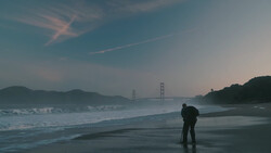 Person standing on wet beach at dusk with suspension bridge visible across the bay.