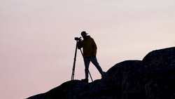 Silhouetted photographer standing on a rocky outcrop while looking through a tripod-mounted camera during twilight.