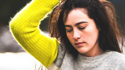 Woman with long dark hair wearing a bright yellow-green cardigan over a gray sweater, looking downward with a contemplative expression.