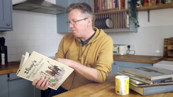 Man in yellow cardigan examining a printed publication at a wooden desk in a kitchen.