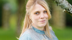 Young woman with blonde hair looking at camera in outdoor setting with green background.