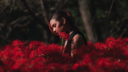 Woman with dark hair and closed eyes surrounded by vibrant red flowers in an outdoor setting.
