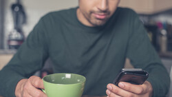 Man holding a green mug and smartphone, looking at phone screen while seated indoors.