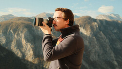 Photographer with glasses holding a DSLR camera with telephoto lens, shooting in a mountainous landscape.